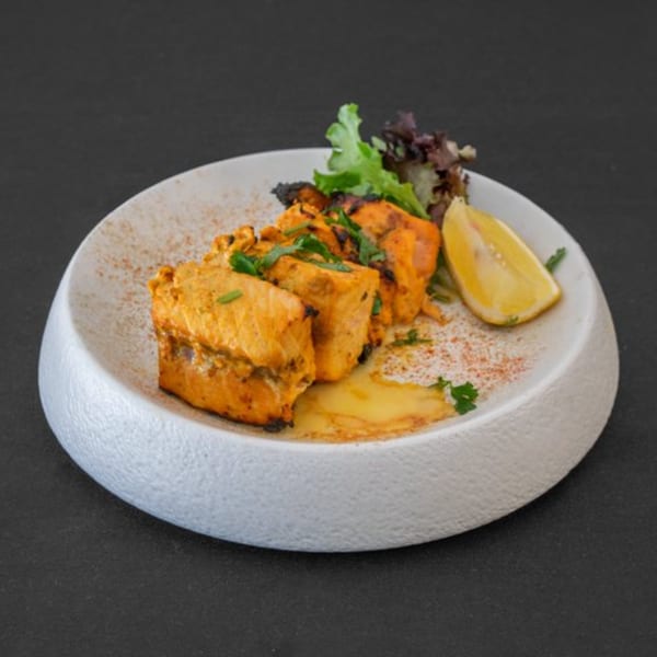 Four pieces of grilled salmon tikka with lemon, lettuce, and sauce on a white plate.