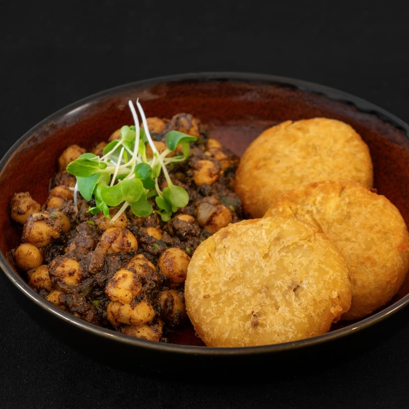 A dark bowl holds spiced chickpeas topped with microgreens, alongside three golden fried flatbreads.