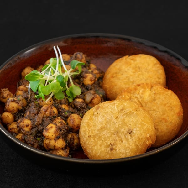 A dark bowl filled with spiced chickpeas and three golden fried bread pieces, garnished with microgreens.