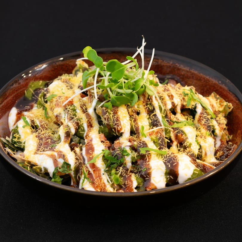 A bowl of Indian chaat, topped with yogurt, chutneys, sev, and microgreens on a black background.