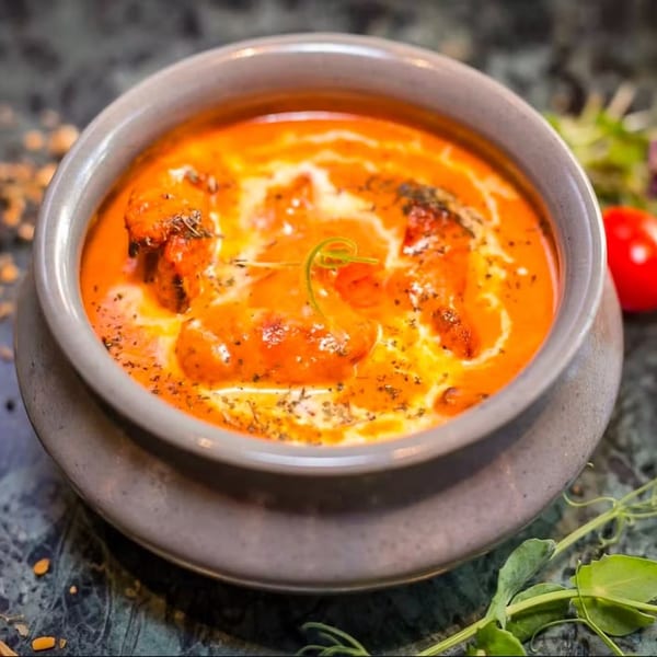 A bowl of vibrant orange butter chicken, garnished with cream swirls and herbs, on a dark marble surface.
