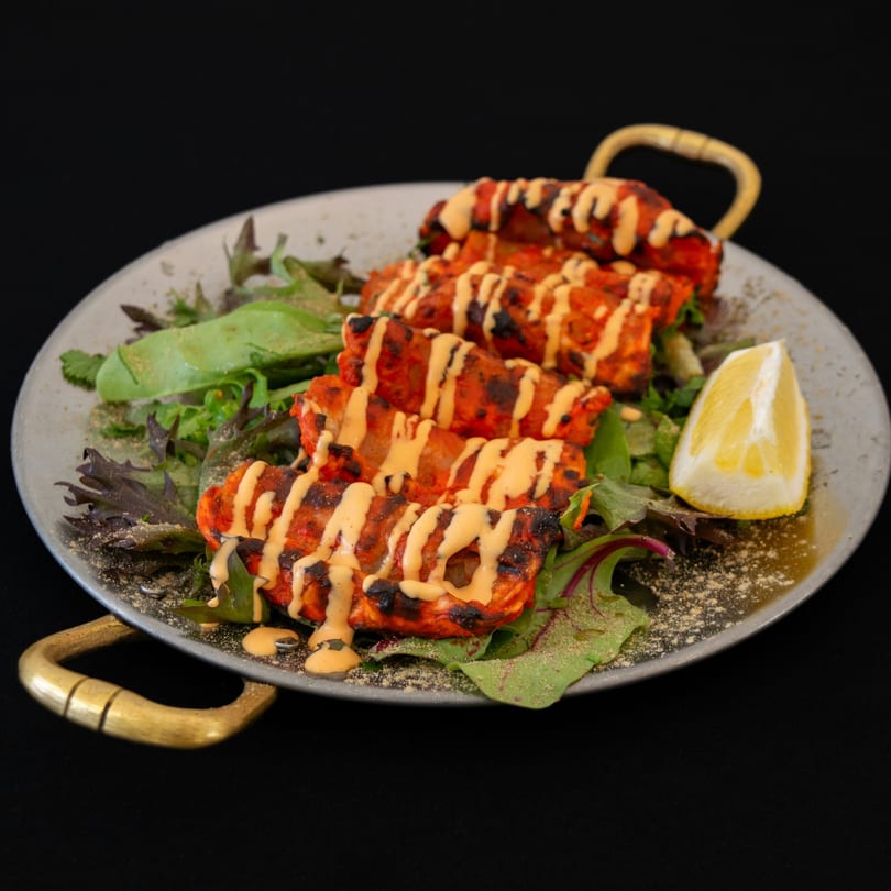 Four grilled chicken skewers with sauce, lettuce, and a lemon wedge on a metal plate.